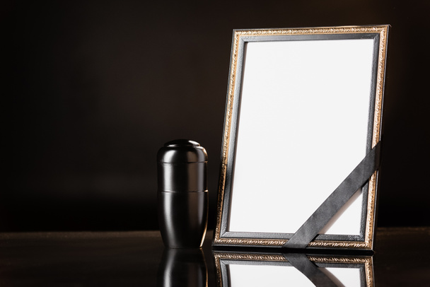 τεφροδόχος με στάχτες, καθρέφτης με μαύρη κορδέλα σε μαύρο φόντο, έννοια κηδεία - Φωτογραφία, εικόνα