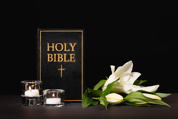 Лили, свечи и священная библия на черном фоне, похоронная концепция - Фото, изображение