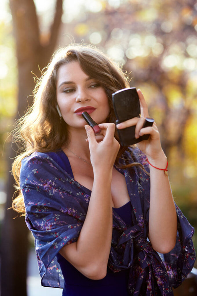 Beautiful attractive woman paints lips with red lipstick in the park. The bright colors of fall - Φωτογραφία, εικόνα