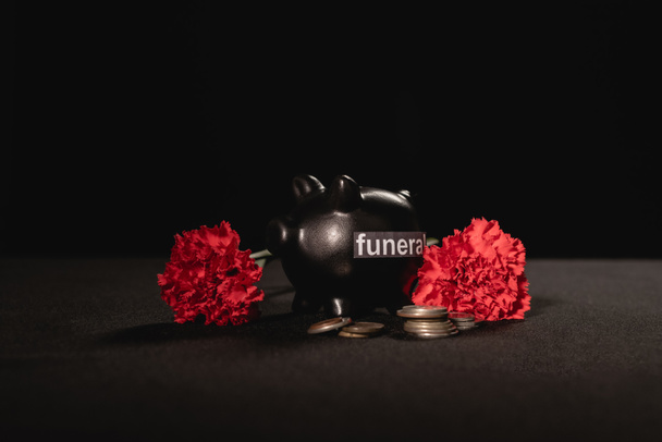 red carnation flowers and piggy bank with coins on black background, funeral concept - Foto, afbeelding