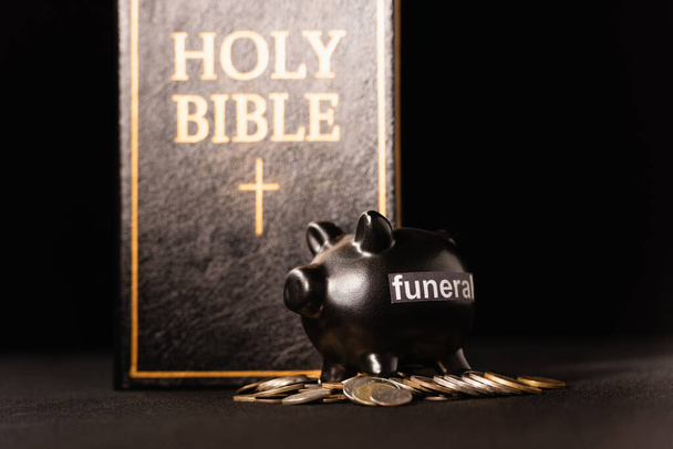 prasečí banka s mincemi a svatou biblí na černém pozadí, pohřební koncept - Fotografie, Obrázek