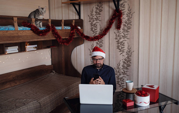 Koyu mavi tişörtlü mutlu bir erkek internetten telefon aracılığıyla alışveriş yapıyor. Yeni yıl için bir sürü manzara. Kırmızı Noel Baba şapkası takan ev sahibi bir işadamından - Fotoğraf, Görsel