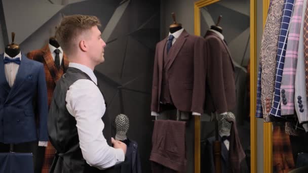 A man puts on a vest. Businessman looks in the mirror on his reflection at the. Businessman looks in the mirror in a luxury men's clothing store against the backdrop of suits, shoes and a mannequin. - Footage, Video