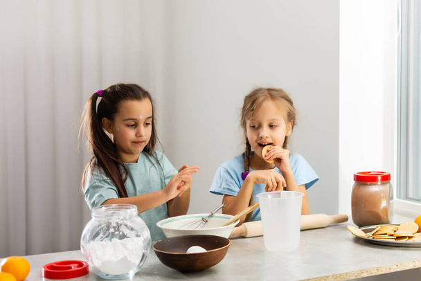 Dwie małe dziewczynki w kuchni przygotowują jedzenie, deser dla rodziny. Kiedy uczą się gotować, zaczynają bawić się mąką i uśmiechać się do siebie. Pojęcie: lekcje gotowania, rodzina, edukacja. - Zdjęcie, obraz