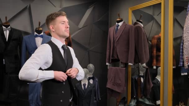 A man puts on a vest. Businessman looks in the mirror on his reflection at the. Businessman looks in the mirror in a luxury men's clothing store against the backdrop of suits, shoes and a mannequin. - Footage, Video