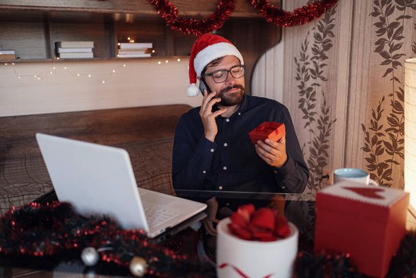 Close up portrait of joyful Caucasian handsome man in santa hat sitting in cozy room xmas decoration calling by smartphone with happy face . New Year. Merry Christmas - Photo, Image
