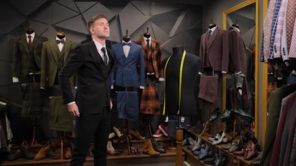 muž si urovná kravatu před zrcadlem v luxusním butikovém obchodě. Portrét muže oblečeného v černém třídílném obleku na pozadí obleků, bot a figuríny. - Záběry, video