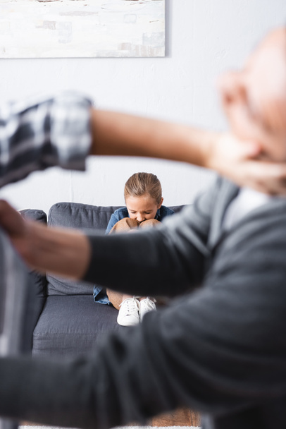 Niño molesto sentado en el sofá mientras los padres pelean en primer plano borroso  - Foto, Imagen