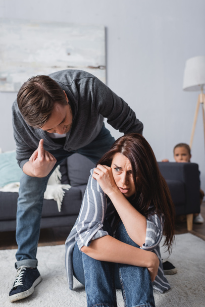 Scared woman sitting on floor near abusive husband pointing with finger and child hiding behind couch on blurred background  - Фото, зображення