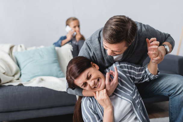 Angry man choking wife while child talking on smartphone on blurred background  - Photo, Image