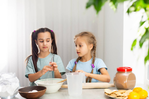 台所の2人の小さな女の子は、家族のためのデザートを準備します。彼らは料理を学ぶと、彼らは小麦粉で遊び始め、お互いに笑顔。概念：料理教室、家庭、教育. - 写真・画像