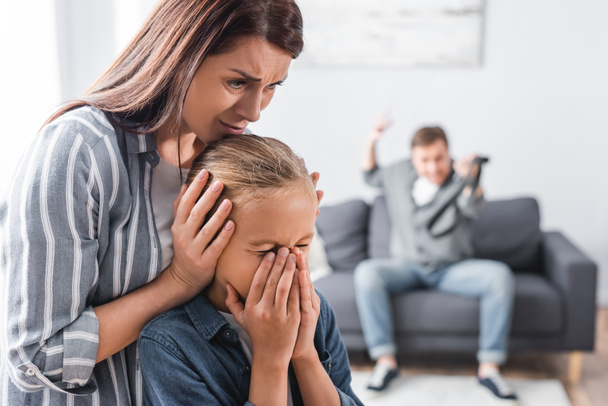 Przestraszona kobieta zasłaniająca uszy córce w pobliżu agresywnego męża na niewyraźnym tle w domu  - Zdjęcie, obraz
