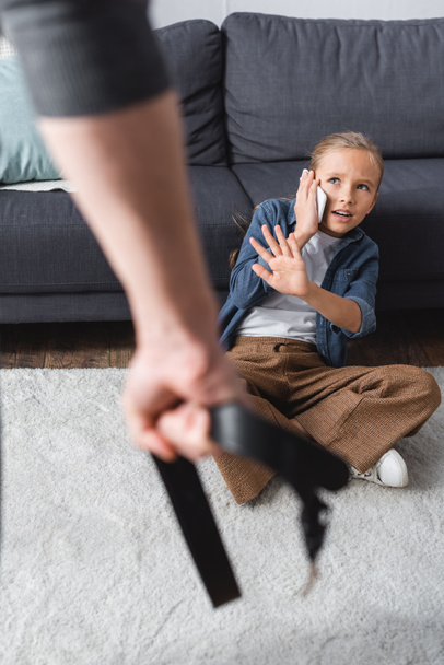 Chica asustada hablando en el teléfono inteligente y mostrando gesto de stop a padre con cinturón en primer plano borroso  - Foto, imagen
