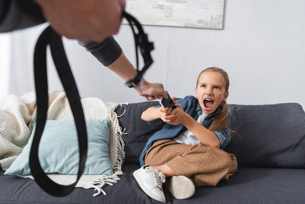 Chico gritando sosteniendo teléfono inteligente cerca del padre con cinturón en primer plano borroso  - Foto, Imagen