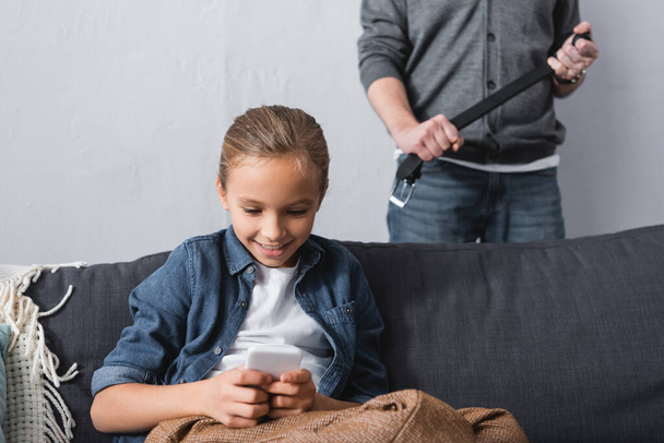 Niño sonriente usando teléfono inteligente cerca del padre con cinturón en el fondo borroso en casa  - Foto, imagen