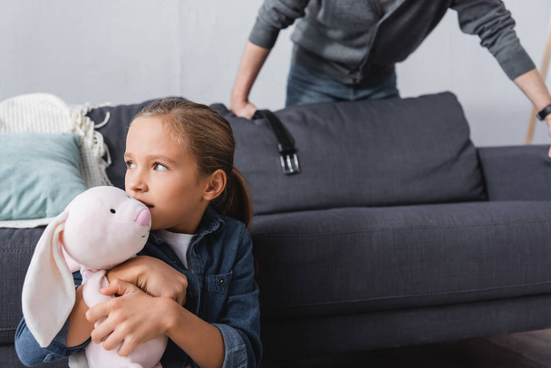 Girl holding soft toy near abusive father with waist belt on blurred background  - Foto, Bild