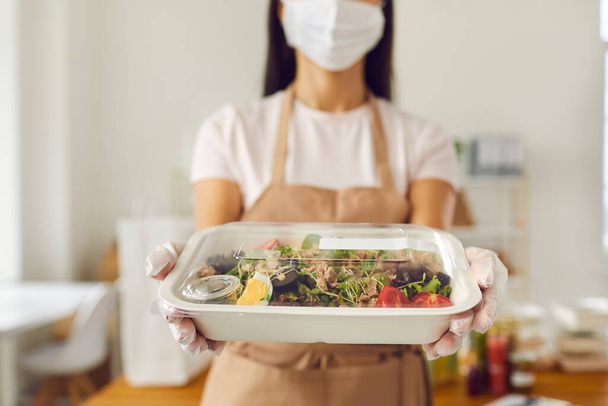 Responsible takeaway cafe worker in face mask holding packed lunch ready for delivery - 写真・画像