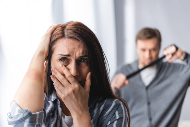 Worried woman with bruise on hand covering mouth near abusive husband with waist belt on blurred background  - Photo, Image