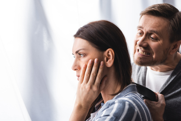 Mujer con la mano cerca de la mejilla mirando al lado de marido irritado con cinturón en casa  - Foto, imagen