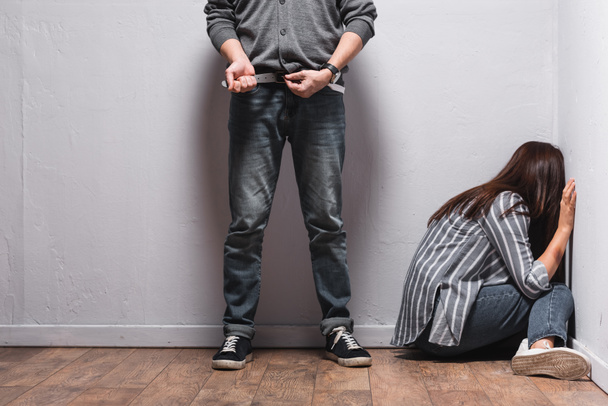 Abusador quitándose el cinturón de cintura cerca de la mujer con moretones en la mano sentado en el suelo junto a las paredes  - Foto, imagen