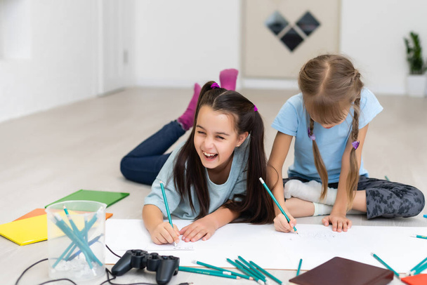 Δύο μικρές αδερφές ξαπλώνουν στο πάτωμα του σπιτιού και ζωγραφίζουν με χρωματιστά μολύβια σε χαρτί. Τα παιδιά κάνουν δημιουργική εργασία μετά το σχολείο. - Φωτογραφία, εικόνα