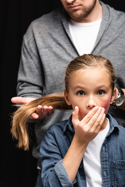 Scared kid with bruise on face standing near abusive father touching hair on blurred background isolated on black  - Photo, Image