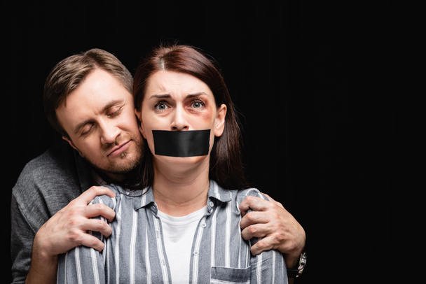 Femme effrayée avec hématome et ruban adhésif sur la bouche debout près de l'agresseur isolé sur noir  - Photo, image
