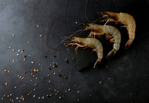 An overhead shot of raw shrimps on a black rock slate with sprinkled rock salt and black pepper - Foto, Imagem