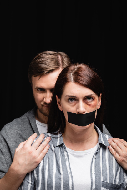 Abuser hiding behind wife with bruise and adhesive tape on mouth isolated on black  - Photo, Image