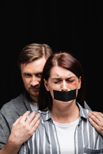 Жестокий муж обнимает депрессивную жену с клейкой лентой и синяком, изолированным на черном  - Фото, изображение