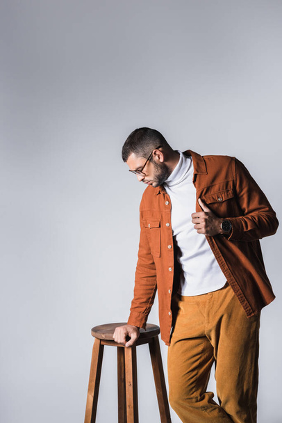 Fashionable man adjusting jacket near wooden chair isolated on grey  - Фото, зображення