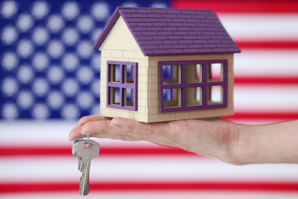 Wooden house and keys are lying on hand in front of US flag closeup - Фото, изображение