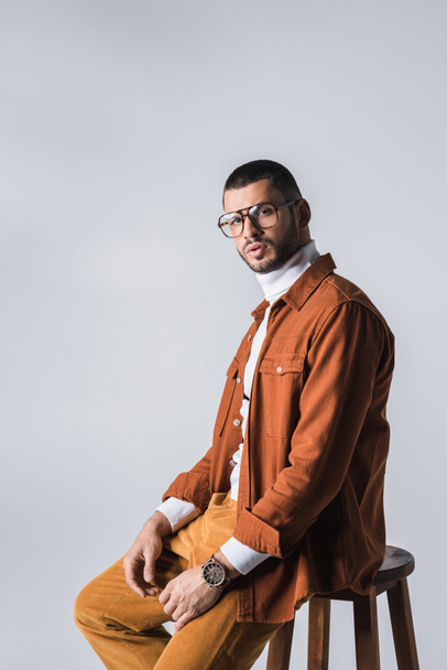 Stylish man in eyeglasses and terracotta jacket sitting on chair isolated on grey  - Foto, imagen