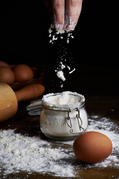 A hand taking flour from a bowl with eggs and a rolling pin on the table - Foto, imagen