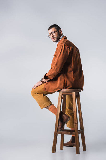 Stylish man in eyeglasses and terracotta jacket looking at camera on wooden chair on grey background  - Foto, afbeelding