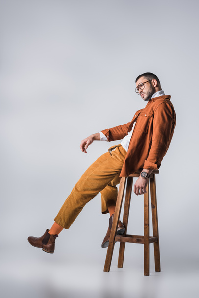 Stylish man in eyeglasses and terracotta jacket posing on wooden chair on grey background  - Photo, image