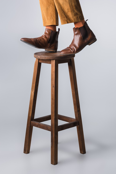 Vista ritagliata dell'uomo in scarpe marroni in piedi su sedia in legno su sfondo grigio  - Foto, immagini