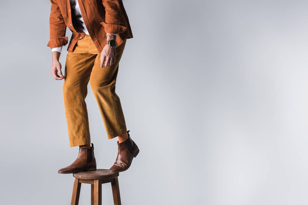 Cropped view of fashionable man in brown shoes standing on wooden chair on grey background  - Photo, Image