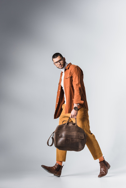 Homme élégant regardant la caméra tout en portant un sac brun et en marchant sur fond gris  - Photo, image