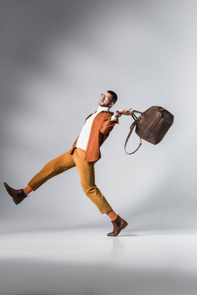 Fashionable man in terracotta jacket holding brown bag while walking on grey background - Foto, Imagem