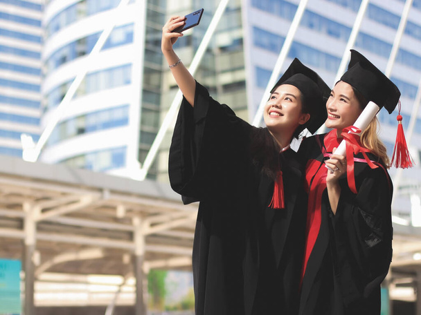 Dos mujeres graduadas felices en vestidos de graduación haciendo foto selfie con teléfono móvil. Edificio de la ciudad. - Foto, Imagen