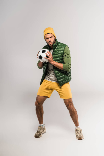 Deportista enfocado en sombrero que sostiene el fútbol sobre fondo gris  - Foto, Imagen