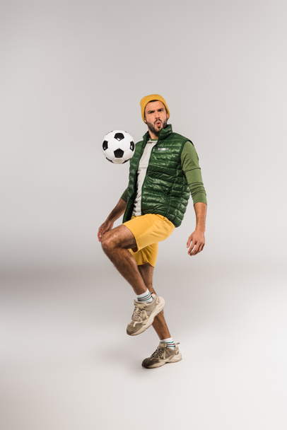 Homem barbudo olhando para a câmera perto de futebol no ar no fundo cinza  - Foto, Imagem