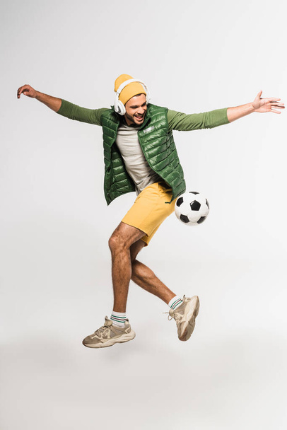 Cheerful sportsman in headphones jumping near football on grey background  - Photo, Image