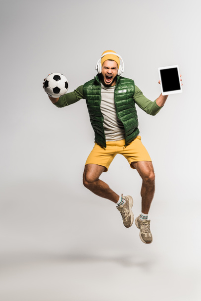 Excited sportsman in headphones holding football and digital tablet with blank screen while jumping on grey background  - Photo, Image
