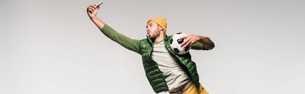 Young sportsman holding football and taking selfie on smartphone isolated on grey, banner  - Foto, Imagem