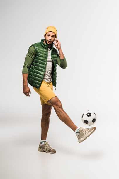 Bearded sportsman talking on smartphone while playing soccer on grey background  - Foto, Imagen