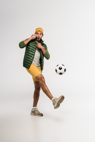 Excited sportsman pointing with finger while talking on smartphone and playing football on grey background  - Фото, изображение