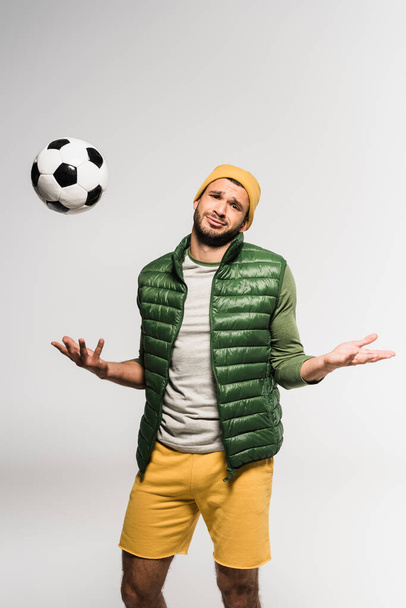 Confused sportsman showing shrug gesture near football in air on grey background  - Фото, изображение