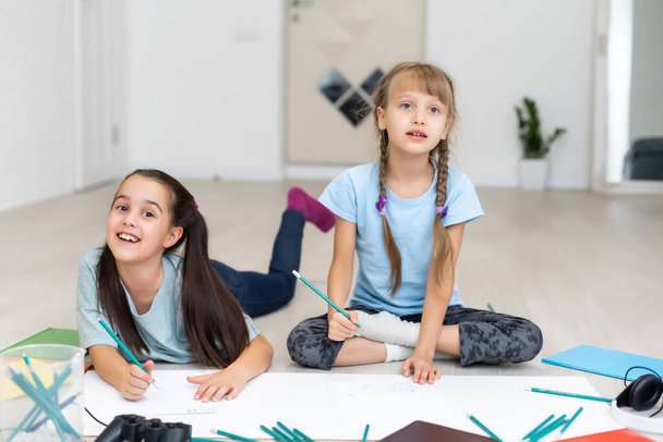 Két kislány nővér fekszik a ház padlóján, és színes ceruzákat rajzolnak papírra. A gyerekek kreatív házi feladatot csinálnak iskola után.. - Fotó, kép